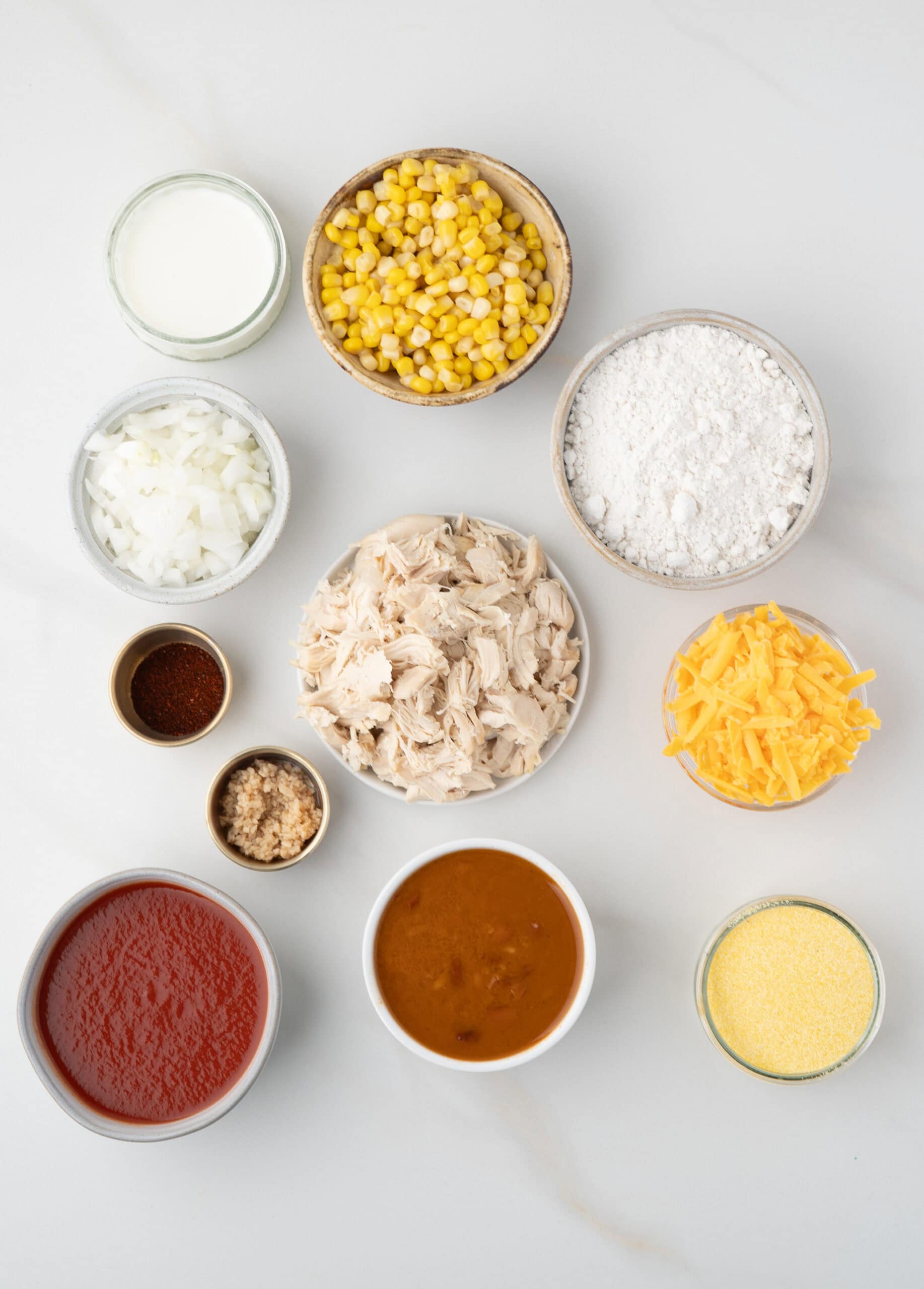 All ingredients to make chicken chili with dumplings: shredded chicken, tomato sauce, corn, onion, garlic, chili powder, Bisquick mix, cornmeal, milk, and cheddar cheese
