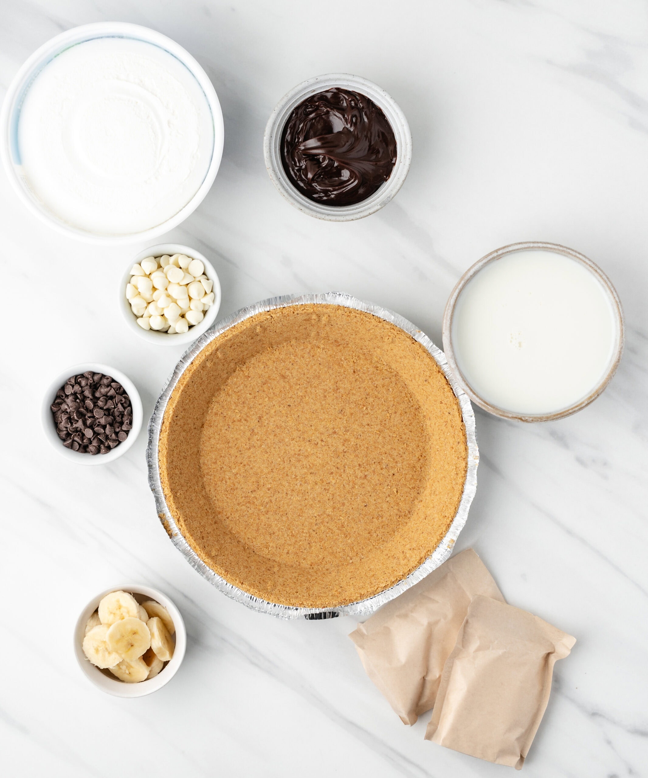 All ingredients to make banana cream pie: graham cracker crust, banana cream pudding mix, milk, hot fudge, white and chocolate chips. sliced bananas, Cool Whip