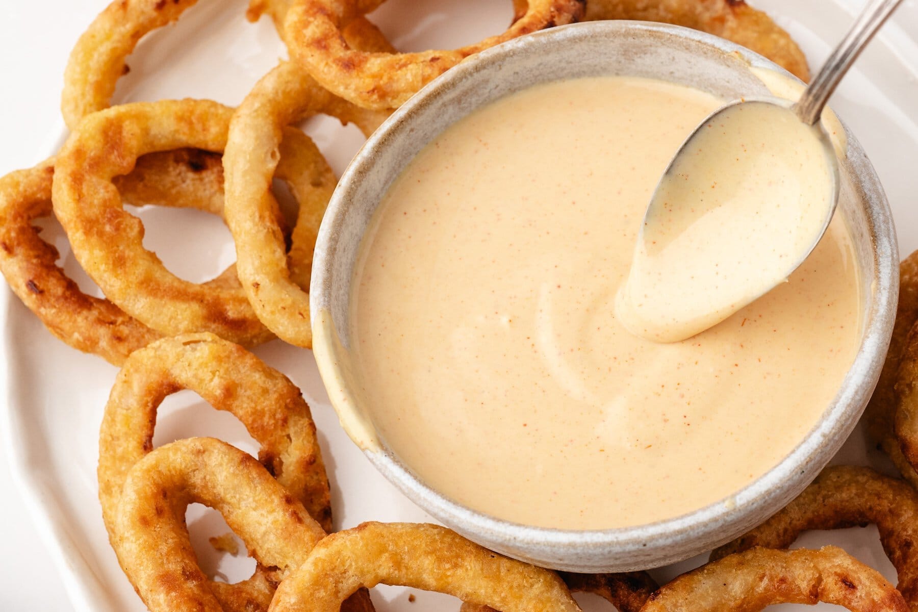 a spoon in a bowl of onion ring dipping sauce surrounded by onion rings