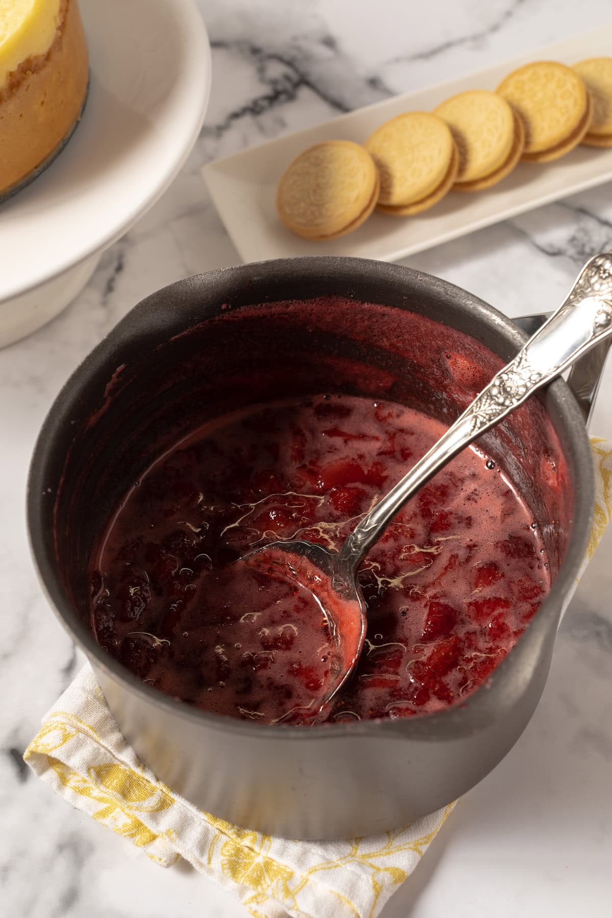 homemade strawberry sauce in a pan with a spoon and a cheesecake in the background